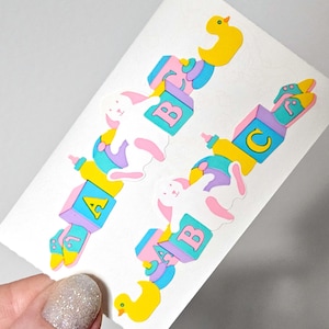 May include: Two decorative baby-themed cutouts on a white background. Each cutout features a baby bottle, a block with a letter, a white bunny, and a yellow duck. The blocks have the letters A, B, and C.