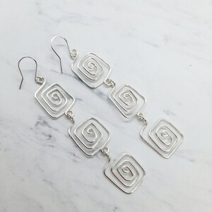 May include: Silver dangle earrings with a geometric design featuring multiple square shapes with a spiral pattern inside each square.