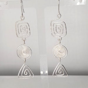 May include: Silver dangle earrings with a geometric design. Each earring features a square, a circle, and a triangle, all made of wire and with a spiral pattern.