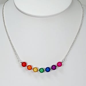 May include: A silver chain necklace with seven rainbow-coloured beads. The beads are red, orange, yellow, green, blue, purple, and pink. The necklace is displayed on a white mannequin.