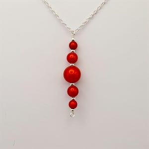 May include: A silver chain necklace with a pendant of red beads. The pendant has a vertical design with varying bead sizes, the largest in the middle. The necklace is set against a white backdrop.
