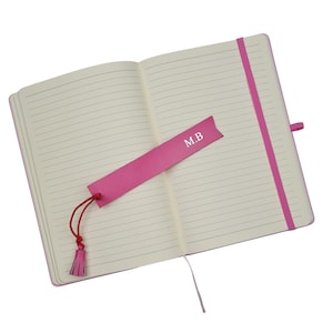 May include: A pink lined notebook with a pink ribbon bookmark. The bookmark has the initials "M.B." printed on it.