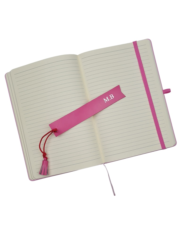 May include: A pink lined notebook with a pink ribbon bookmark. The bookmark has the initials "M.B." printed on it.