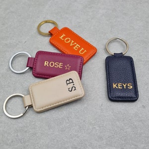May include: Four rectangular leather keyrings in orange, burgundy, beige, and navy blue. The orange keyring says "LOVE U", the burgundy keyring says "ROSE", the beige keyring has "S.B", and the navy blue keyring says "KEYS". Each keyring has a metal ring.