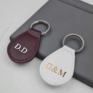 Könnte beinhalten: Zwei Leder-Schlüsselanhänger mit Metallringen. Einer ist burgunderfarben mit weißen Buchstaben, die "D.D." sagen, und der andere ist weiß mit goldenen Buchstaben, die "G&M." sagen.