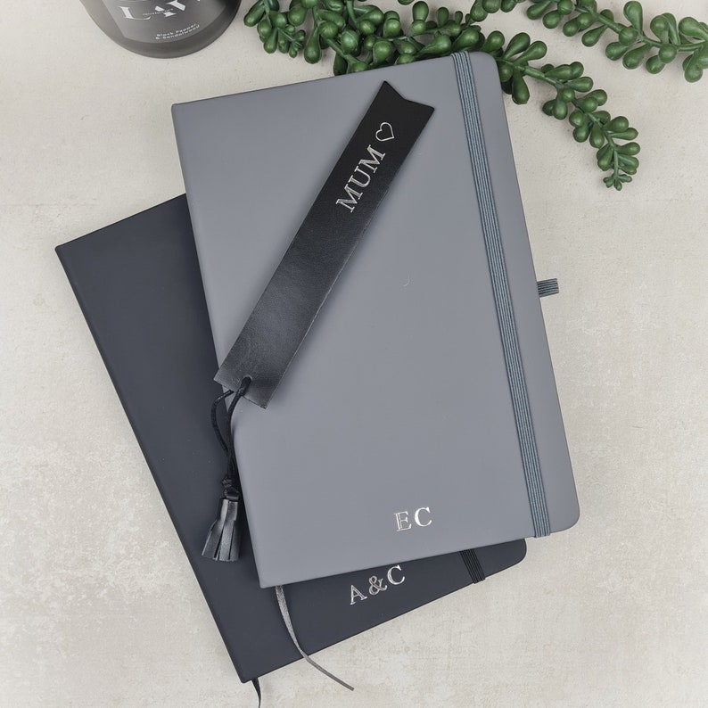 May include: Two gray hardcover journals with black leather bookmarks. The top journal has the word "MUM" with a heart symbol on the bookmark. The bottom journal has the initials "A&C" and "EC" on the covers.
