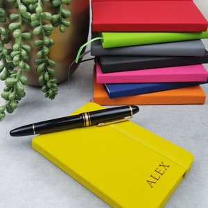 Puede incluir: Una libreta amarilla con el nombre "ALEX" grabado en la portada. Un bolígrafo negro y dorado está sobre la libreta. Hay una pila de libretas de colores en el fondo.