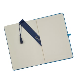 May include: An open blue notebook with a blank page and a blue bookmark with the initials "JJ" and a heart. The bookmark has a tassel.