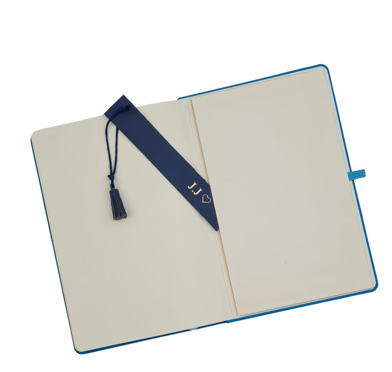 May include: An open blue notebook with a blank page and a blue bookmark with the initials "JJ" and a heart. The bookmark has a tassel.