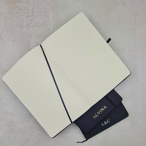 Może przedstawiać: Czarny notes z czarną wstążką zamykającą i napisem "ALVINA" nadrukowanym złotą czcionką na okładce. Notes jest otwarty, a wewnętrzne strony są puste. Notes leży na mniejszym czarnym notesie z literami "A & C" nadrukowanymi złotą czcionką na okładce.