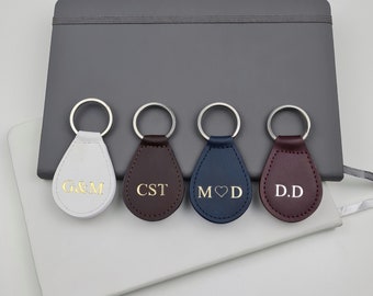 May include: Four leather teardrop-shaped keychains in white, brown, navy, and burgundy. Each keychain has a silver ring and gold lettering: G&M, CST, M with a heart and D, and D.D. A gray notebook and a white notepad are in the background.