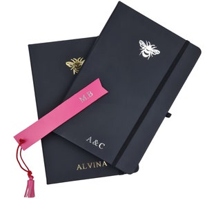 May include: Two black notebooks with gold and silver bee accents. The top notebook has a silver bee and the text "A & C ALVINA". The bottom notebook has a gold bee and a pink bookmark with the text "M.B."