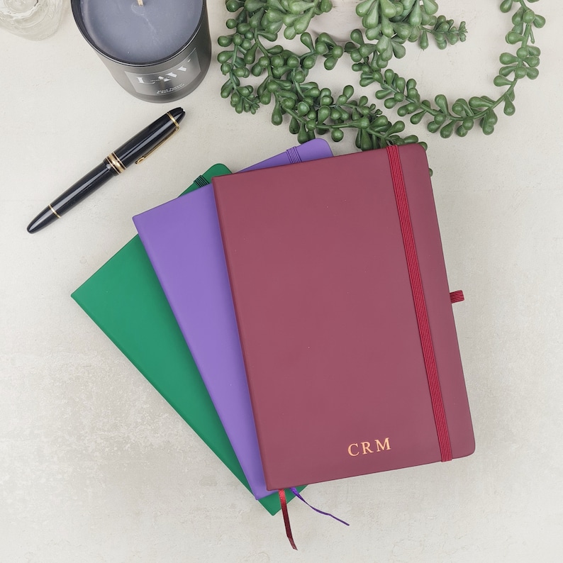 May include: Three notebooks stacked on top of each other. The top notebook is burgundy with a gold ribbon and the text "CRM" on the front. The middle notebook is purple and the bottom notebook is green.