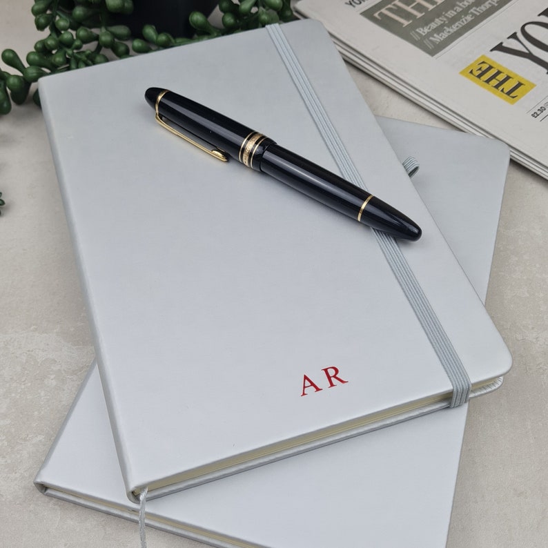 May include: Two silver notebooks with a black pen on top of the top notebook. The top notebook has the initials "AR" printed in red on the front cover.