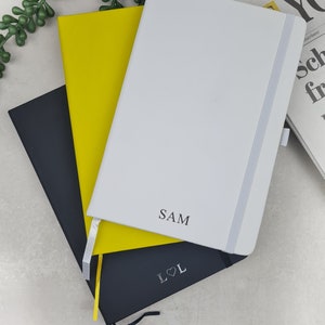 May include: Three notebooks stacked on top of each other. The top notebook is white with a black inscription that says "SAM". The middle notebook is yellow. The bottom notebook is black with a silver inscription that says "L&hearts;L".