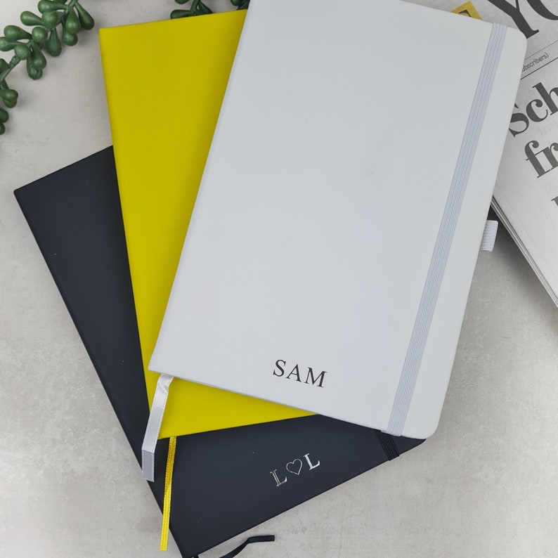 May include: Three notebooks stacked on top of each other. The top notebook is white with a black inscription that says "SAM". The middle notebook is yellow. The bottom notebook is black with a silver inscription that says "L&hearts;L".