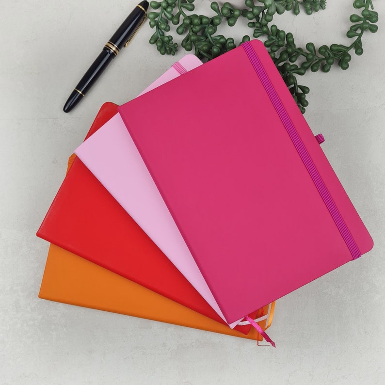 May include: Four notebooks stacked on top of each other. The top notebook is pink with a pink elastic band. The second notebook is light pink. The third notebook is orange. The bottom notebook is red. All notebooks have a ribbon bookmark.