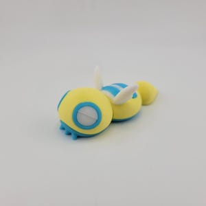 May include: A pair of whimsical, yellow and blue toy figures with white wing-like appendages. The figures have a spherical body with blue accents and a circular blue and white design. The toys are designed to resemble a bee.