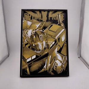 Op de afbeelding: Een rechthoekig ingelijst wandkunstwerk met een gestileerde illustratie van Optimus Prime in goud-, zwart- en wittinten. De tekst OPTIMUS PRIME staat in een vetgedrukt bloklettertype bovenaan het kunstwerk, dat is gevat in een zwarte lijst.