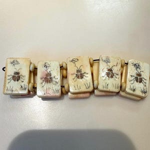 May include: Beige rectangular bracelet with a black string. Each rectangle features a hand-drawn beetle illustration with grass and a small insect. The bracelet is made of rectangular and cylindrical beads.