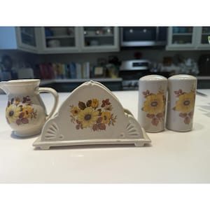 May include: A vintage cream-colored ceramic dining set featuring a creamer pitcher, napkin holder, and salt and pepper shakers. Each piece is decorated with a yellow sunflower and brown leaf design.