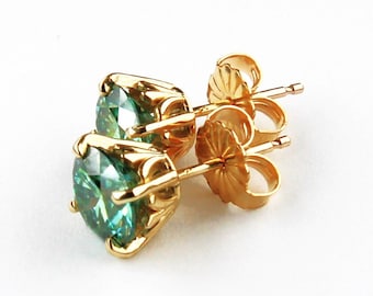 May include: A pair of gold-colored stud earrings with round, teal-colored gemstones. The earrings are set in a four-prong setting, and the posts and backs are also gold-colored. The earrings are displayed on a white background.