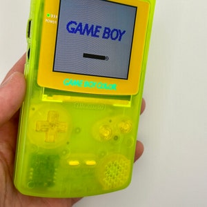 Puede incluir: Una consola de videojuegos portátil Game Boy Color de color amarillo brillante con una pantalla amarilla y negra. La pantalla muestra el texto "GAME BOY" y "GAME BOY COLOR".