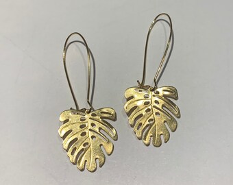 May include: A pair of gold-tone earrings featuring a cutout monstera leaf design. The earrings are made of metal and have a simple, elegant style.