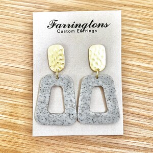 May include: A pair of gray and gold earrings with a hammered gold top and a gray speckled bottom. The earrings are shaped like a rectangle with a smaller rectangle cut out of the center. The earrings are displayed on a white card with the text "Farringtons Custom Earrings".
