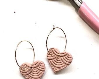 Op de afbeelding: Een paar roze hartvormige oorbellen met een gouden hoepelontwerp. De oorbellen hebben een textuurpatroon en zijn gemaakt van polymer clay.