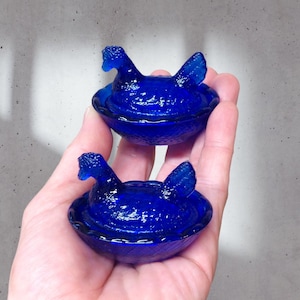 May include: Two cobalt blue glass hen-shaped dish sets. Each set features a hen sitting on a nest.