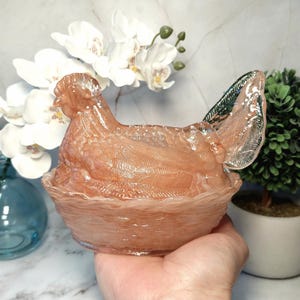 May include: A translucent peach-colored hen-shaped decorative container. The hen is sitting in a nest-like bowl. The hen's wing has a green and silver accent. The container is likely made of glass or a similar material.