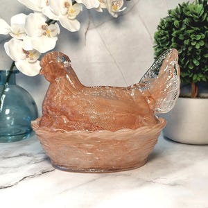 May include: A decorative glass hen figurine in a nest-shaped bowl. The hen is a translucent peach color, with a detailed wing and head. The nest is a matching peach color with a woven texture. The background includes white flowers and a small green plant.