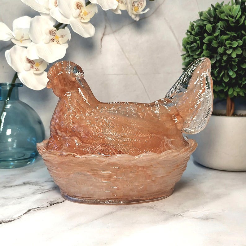 May include: A decorative glass hen figurine in a nest-shaped bowl. The hen is a translucent peach color, with a detailed wing and head. The nest is a matching peach color with a woven texture. The background includes white flowers and a small green plant.
