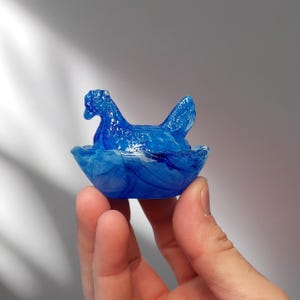 May include: A translucent blue hen figurine in a nest-like bowl. The hen has a detailed head and wing, and the bowl has a swirling pattern. The figurine is held in a hand.