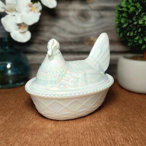 May include: A white iridescent glass hen on a nest dish. The lid is shaped like a sitting hen with detailed feather textures, while the base features a raised diamond lattice pattern and a rope-style rim. The glass has a pearlescent finish.