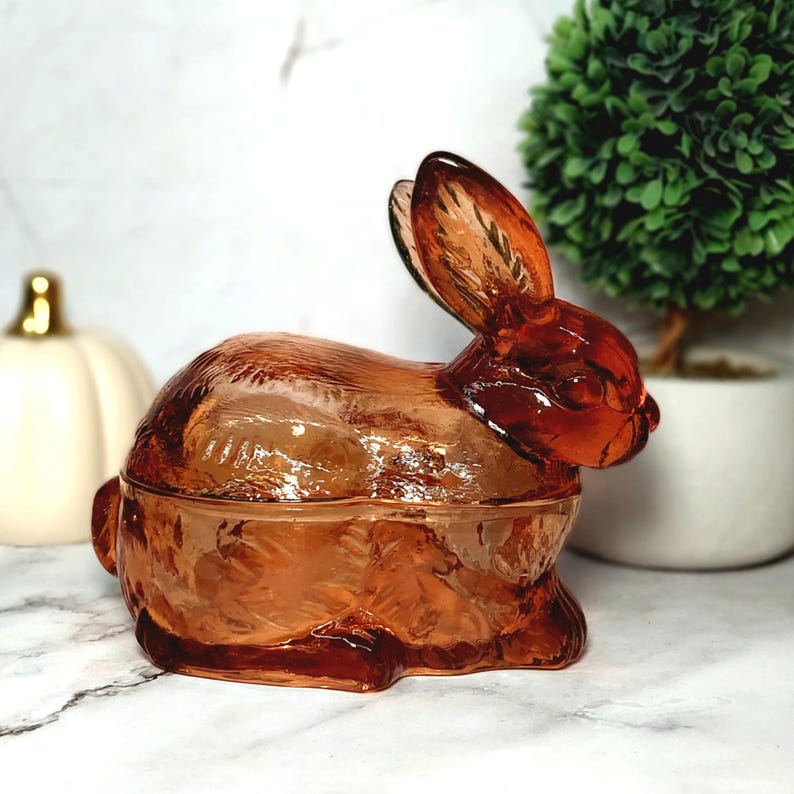Op de afbeelding: Een decoratief amberkleurig glazen konijnbeeldje met een deksel. Het konijn zit met opgerichte oren. Het glas heeft een getextureerd oppervlak en is doorschijnend. Een charmant decoratief item met Pasen- of lentethema.