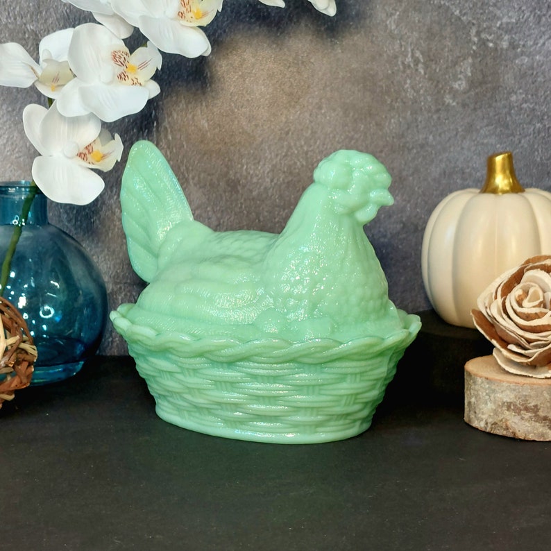 May include: A light green hen-shaped trinket box. The hen sits atop a woven basket base. The box is made of glass or ceramic. The background includes white flowers, a blue vase, and a white pumpkin.