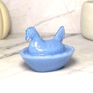 May include: A light blue hen figurine in a basket-shaped bowl. The hen has a detailed comb and wattle, with its wings slightly raised. The bowl has a textured, woven pattern. The figurine is made of a translucent material.