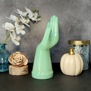 May include: A light green ceramic hand sculpture with a raised finger, sitting on a black surface with a white pumpkin, a wooden log, and a clear glass vase with a floral arrangement.