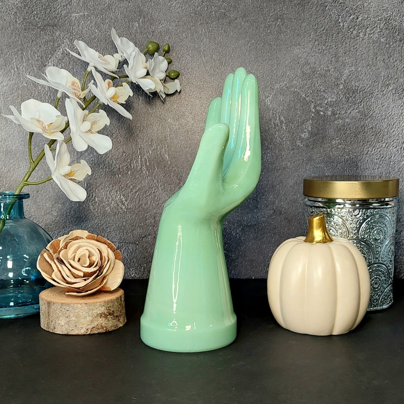 May include: A light green ceramic hand sculpture with a raised finger, sitting on a black surface with a white pumpkin, a wooden log, and a clear glass vase with a floral arrangement.