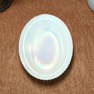 May include: An oval-shaped white milk glass dish featuring a beaded rim and a central iridescent finish that displays subtle rainbow-like reflections. The bowl has a smooth, glossy surface and a distinct, raised decorative border around the edge.