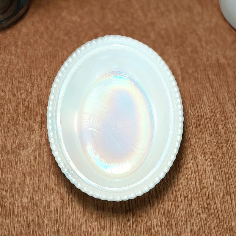 May include: An oval-shaped white milk glass dish featuring a beaded rim and a central iridescent finish that displays subtle rainbow-like reflections. The bowl has a smooth, glossy surface and a distinct, raised decorative border around the edge.
