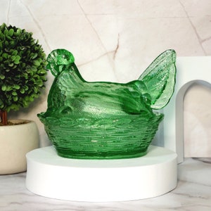 May include: A green glass hen-shaped dish with a basket weave base. The hen is perched atop the basket, with detailed feather and wing textures. The dish is a vibrant green color and sits on a white pedestal.