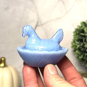 May include: A light blue glass hen figurine in a basket-like bowl. The hen is sitting in the bowl, with its head and tail visible. The bowl has a textured, woven pattern. The figurine is held in a hand.