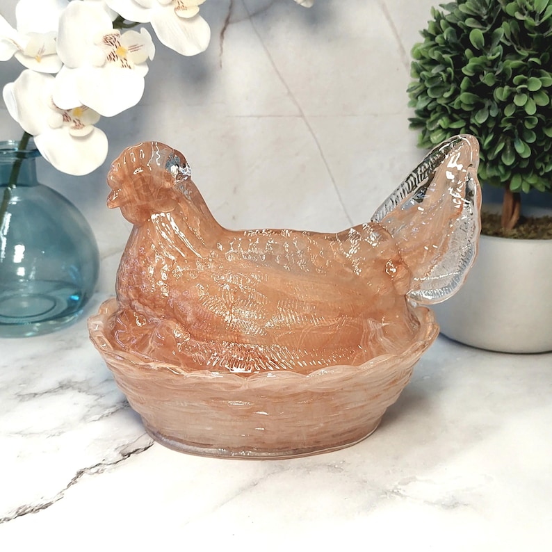 May include: A decorative, translucent hen figurine in a nest shape. The hen is a light peach color with a textured surface, and the nest has a scalloped edge. The figurine is likely made of glass or a similar material.