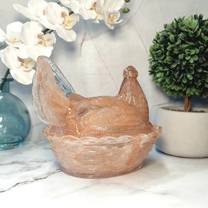 May include: A decorative glass hen-shaped container in shades of peach and light brown. The hen sits atop a basket-like base. The background includes white orchids, a blue vase, and a small green topiary.