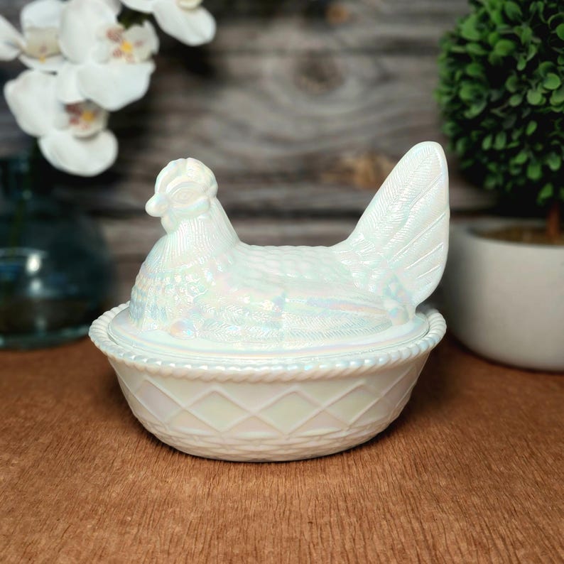 May include: An iridescent white glass hen on a nest dish. The lid is shaped like a sitting hen with detailed feather textures, while the base features a raised diamond lattice pattern and a rope-style rim, creating a glossy, pearlescent finish.