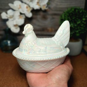 May include: A white iridescent glass hen on a nest dish. The lid is shaped like a sitting hen with detailed feathers, while the base features a textured basket-weave pattern with a beaded rim. The glass has a pearlescent finish that reflects light.