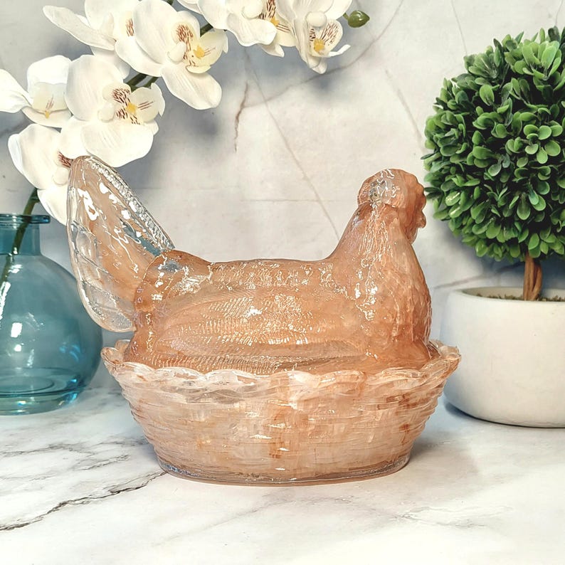 May include: A decorative glass hen-shaped container in a basket weave design. The hen is a translucent peach color, and the basket is a similar shade. The container is likely used for decorative storage or display.